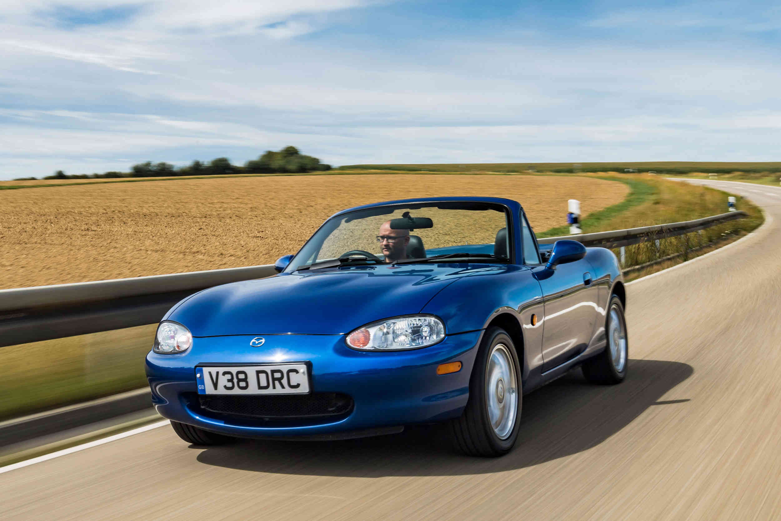 A Mazda MX-R with open top driving down a road through the country